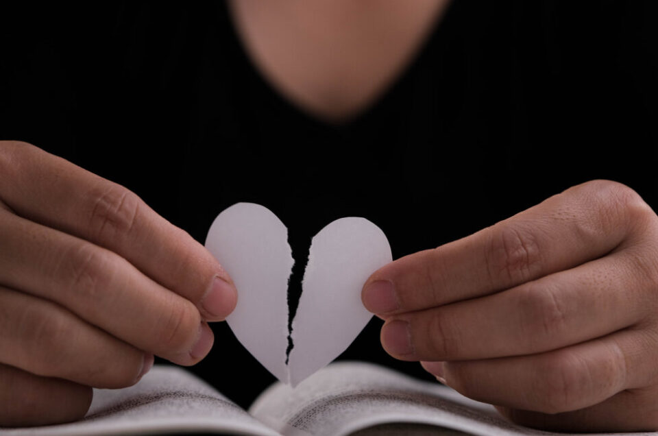 Female hand holding a half-torn white paper heart over an open book on the table. White paper heart torn in half in hand